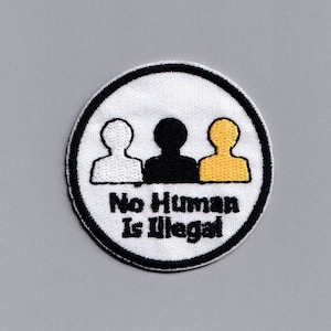 Könnte beinhalten: Ein weißer, schwarzer und gelber gestickter Aufnäher mit drei Strichmännchen, die verschiedene Hautfarben darstellen. Der Text "No Human Is Illegal" ist in Schwarz unter den Figuren geschrieben.