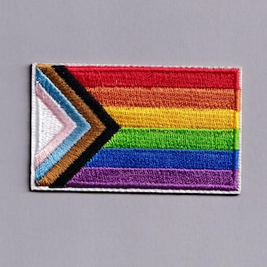 Può includere: Una toppa rettangolare ricamata con il design della bandiera Progress Pride. La toppa presenta strisce orizzontali rosse, arancioni, gialle, verdi, blu e viola, con uno chevron nero a sinistra contenente strisce azzurre, rosa e marroni.