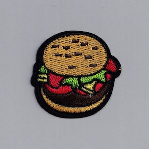 Pode incluir: Um remendo bordado de um hambúrguer de desenho animado com alface, tomate e queijo, em marrom e amarelo.