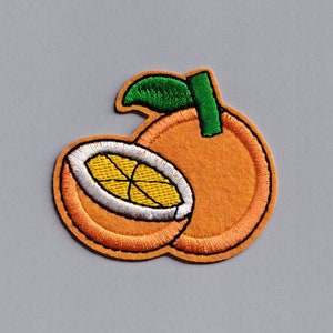 Puede incluir: Un parche bordado de una naranja con una hoja verde, una naranja está entera y la otra está cortada por la mitad, mostrando el interior de la fruta.