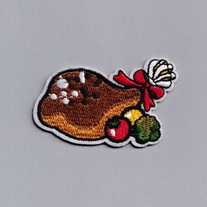 May include: An embroidered iron-on patch featuring a cartoon illustration of a roasted turkey leg with a red ribbon and a white flower. The turkey leg is surrounded by a green sprig of parsley, a red tomato, and a yellow onion.