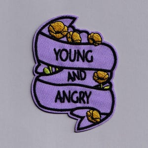 May include: Embroidered patch in a purple banner shape with the words "YOUNG AND ANGRY" in black. The patch features yellow flower accents and a black border.