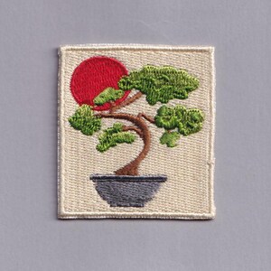 May include: Embroidered patch depicting a bonsai tree in a grey pot with a red sun. The tree has green leaves and a brown trunk. The patch is rectangular with a beige background and a stitched border.