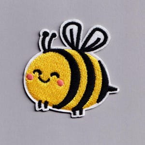 Può includere: Toppa ricamata raffigurante un'ape dei cartoni animati sorridente. L'ape ha un corpo giallo con strisce nere, guance rosa e antenne e ali nere. La toppa è bordata di bianco.