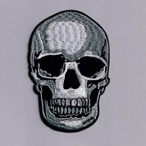 May include: A monochrome embroidered skull patch. The design features a detailed skull with black eye sockets and a defined jaw. The patch is suitable for application on clothing or accessories.