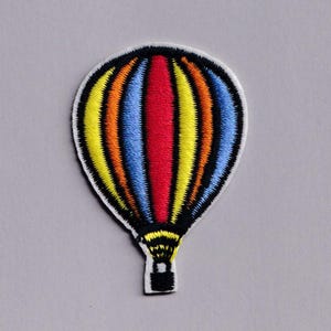 May include: Embroidered patch featuring a colorful hot air balloon design. The balloon has vertical stripes in red, yellow, orange, and blue, outlined in black. The patch has a white border and a black basket.