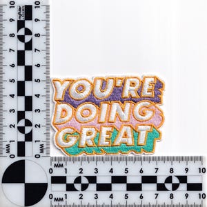 May include: An embroidered iron-on patch with the text "YOU'RE DOING GREAT" in a colorful, layered font style. The words are outlined in yellow and the background is a gradient of purple, pink, and teal.