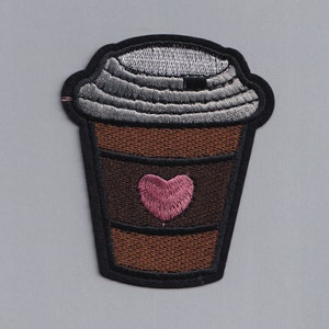 Puede incluir: Parche bordado para planchar de una taza de café marrón con un corazón rosa en la parte delantera. La taza tiene un contorno negro y una tapa gris.