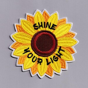Könnte beinhalten: Gestickter Sonnenblumen-Aufnäher mit gelben, orangefarbenen und braunen Blütenblättern. Der Aufnäher trägt die Worte "SHINE YOUR LIGHT" in schwarzer Schrift. Der Aufnäher ist mit weißen Nähten umrandet.