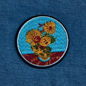 May include: Embroidered patch featuring a stylized depiction of sunflowers in a vase. The sunflowers are yellow with brown centers and green leaves. The vase is brown and white with a blue background. The patch is circular and has a black border.