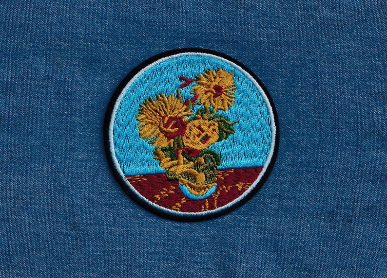 May include: Embroidered patch featuring a stylized depiction of sunflowers in a vase. The sunflowers are yellow with brown centers and green leaves. The vase is brown and white with a blue background. The patch is circular and has a black border.