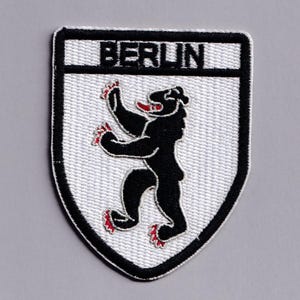Puede incluir: Un parche bordado en forma de escudo con fondo blanco y borde negro. La palabra BERLIN está bordada en negro en la parte superior. Un oso negro con garras y lengua rojas está centrado en el parche en posición erguida.