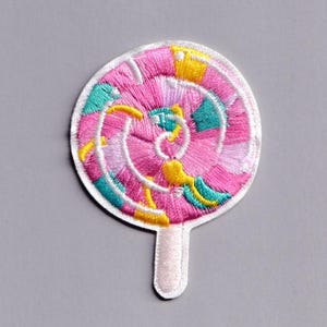 May include: Embroidered patch in the shape of a lollipop. The lollipop features a pink center with a spiral design, accented with teal, yellow, and white details. The patch has a white border and a white stick.