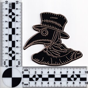 May include: Black and beige embroidered patch of a plague doctor wearing a long coat and a hat with a beak-shaped mask.