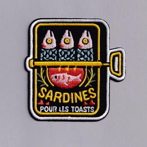May include: Embroidered patch with a sardine can design. The black patch has a yellow border and shows three fish above a red tomato with a fish illustration. The text "SARDINES POUR LES TOASTS" is below the tomato.
