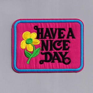 Puede incluir: Un parche de tela rosa con ribete azul, que presenta una flor amarilla con hojas verdes y el texto "HAVE A NICE DAY" en negro.