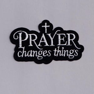 May include: Black embroidered patch with the words "PRAYER changes things" in white script. A white cross is above the word "PRAYER". The patch has a black border and is set against a light grey background.
