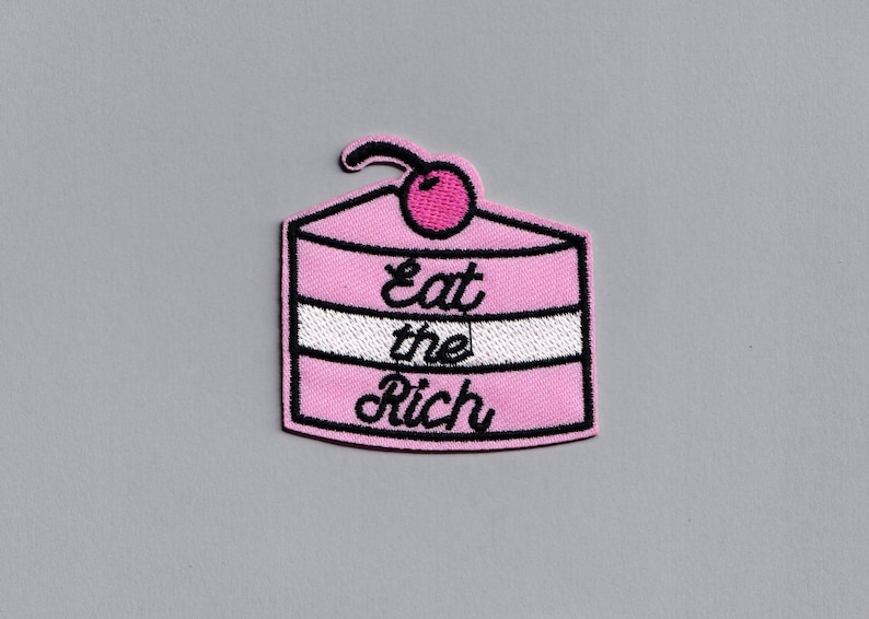 May include: Pink embroidered iron-on patch shaped like a cake with a cherry on top. The cake has white frosting and the text "Eat the Rich" in black.