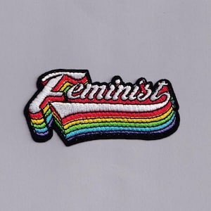 Può includere: Toppa ricamata con la parola "Feminist" in corsivo bianco su un design arcobaleno. La toppa ha un bordo nero ed è su uno sfondo grigio. L'arcobaleno comprende rosso, arancione, giallo, verde, blu e viola.