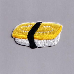 May include: Embroidered sushi patch featuring a yellow topping, white rice, and a black seaweed strip. The patch is designed with detailed stitching and a textured appearance, perfect for adding to clothing or accessories.