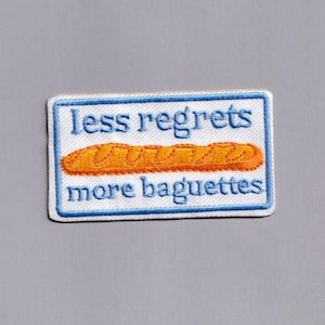 Op de afbeelding: Geborduurde patch met een witte achtergrond en een blauwe rand. De patch heeft de tekst "less regrets more baguettes" in blauw, met een oranje baguette-illustratie in het midden.