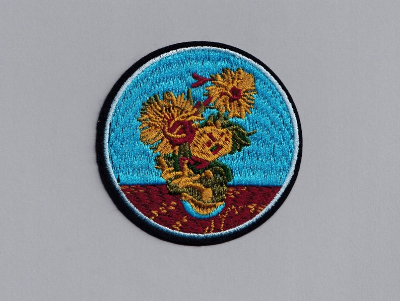 May include: A round embroidered patch with a blue background and a vase of yellow sunflowers. The vase is brown and has a red base. The sunflowers are in a vase with a red base.