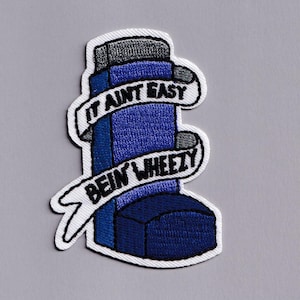 May include: Embroidered patch depicting a blue inhaler with the text "IT AINT EASY BEIN' WHEEZY" on a white banner. The patch has a white border and is set against a grey background.