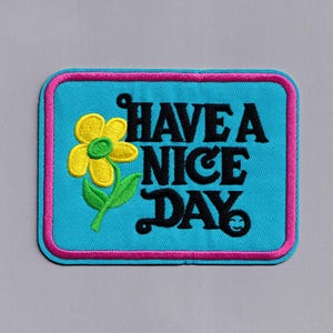 Könnte beinhalten: Ein blauer gestickter Aufnäher mit rosa Nähten. Er zeigt eine gelbe Blume mit einem grünen Stiel und den Text "HAVE A NICE DAY".