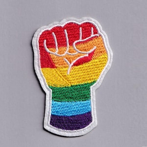 May include: A rainbow-coloured embroidered patch depicting a raised fist, a symbol of solidarity and LGBTQ+ pride. The patch has a white border and is set against a grey background. The fist is made up of red, orange, yellow, green, blue, and purple.