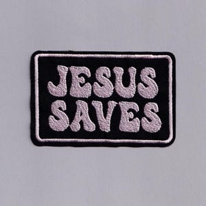 Könnte beinhalten: Ein rechteckiger schwarzer Aufnäher mit den Worten "JESUS SAVES" in hellrosa gestickt, weiß umrandet. Der Aufnäher ist ein religiöser Artikel, der sich zum Aufnähen auf Kleidung oder Accessoires eignet.