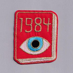 Puede incluir: Parche rojo bordado con el número "1984" en hilo dorado sobre un ojo azul. El parche tiene un borde dorado y un contorno blanco, sobre un fondo gris.