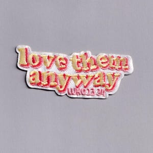 Puede incluir: Parche bordado con la frase "love them anyway" en fuente cursiva. El texto es en hilo amarillo y rojo, con un contorno rojo. El parche tiene un borde blanco y está sobre un fondo gris.