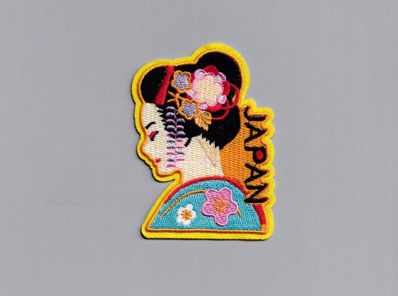 May include: Embroidered patch featuring a geisha in a blue and yellow kimono with pink flowers. The word "JAPAN" is embroidered vertically on the right side of the patch.