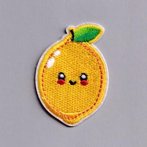May include: Embroidered patch of a smiling lemon with a green leaf. The lemon is yellow with a white outline and red cheeks. The patch is on a gray background.