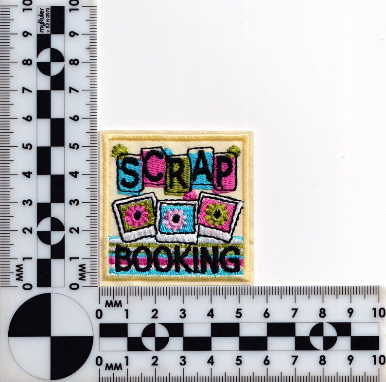 May include: A yellow embroidered patch with the words "SCRAP BOOKING" in colorful letters. The patch features three colorful photo frames with flowers and a blue and green striped background.