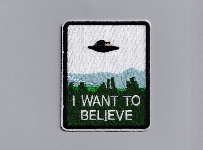 May include: A black and green embroidered patch with a white background. The patch features a flying saucer over a green field with trees and the text "I WANT TO BELIEVE".