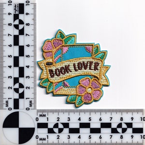 May include: An embroidered iron-on patch with a blue book, a gold banner with the text "BOOK LOVER", and pink and green flowers.