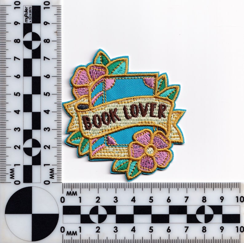 May include: An embroidered iron-on patch with a blue book, a gold banner with the text "BOOK LOVER", and pink and green flowers.