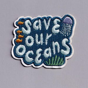 May include: Embroidered patch with the words "Save Our Oceans" in white, outlined in blue. The patch features orange fish, a purple jellyfish, and green seaweed. The patch has a white border.