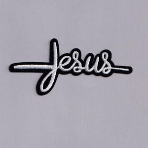 May include: Embroidered patch featuring the word "Jesus" in white script with a black border. The design includes a cross integrated into the letter "J". The patch is likely for clothing or accessories.