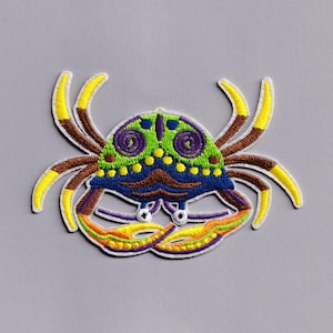 May include: A vibrant crab patch with a decorative design. The crab has a green, blue, and purple body with yellow and brown legs. The patch is outlined in white and has a playful, cartoonish style.