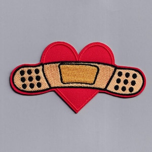 May include: Red felt heart with a brown and beige band-aid over it. The band-aid has a yellow rectangle in the center.