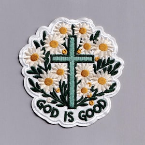 Może przedstawiać: Haftowana naszywka z turkusowym krzyżem otoczonym białymi stokrotkami z żółtymi środkami i zielonymi liśćmi. Naszywka zawiera napis "GOD IS GOOD" w kolorze białym, z białym, falistym obramowaniem. Kwiatowy i religijny.