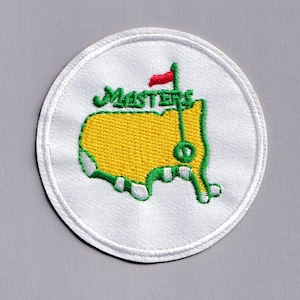 May include: A circular white embroidered patch featuring the Masters Tournament logo. The logo includes a yellow outline of a map with green accents, the word "MASTERS" in green script, and a red flag on a green pole.