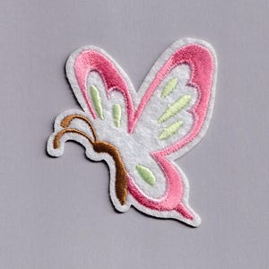 Puede incluir: Parche de mariposa bordada con cuerpo blanco, contornos de alas rosas y detalles en verde claro. La mariposa tiene antenas marrones y está sobre un fondo gris. Ideal para ropa, bolsos u otros artículos de tela.
