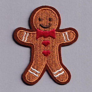 May include: Embroidered gingerbread man patch. It has a brown outline, tan body, and a red bow tie. The patch features white accents on the arms and legs, and two red heart-shaped buttons. The gingerbread man has a smiling face.