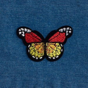 Puede incluir: Un parche bordado de mariposa rojo, amarillo y negro. La mariposa est&aacute; sobre un fondo de mezclilla azul.