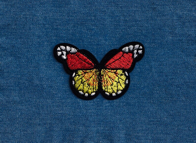 Puede incluir: Un parche bordado de mariposa rojo, amarillo y negro. La mariposa est&aacute; sobre un fondo de mezclilla azul.