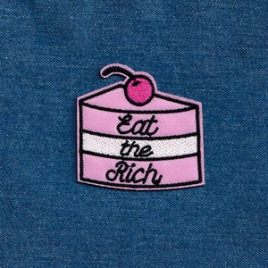 May include: Pink and white embroidered patch of a layered cake with a cherry on top. The text "Eat the Rich" is embroidered on the cake.