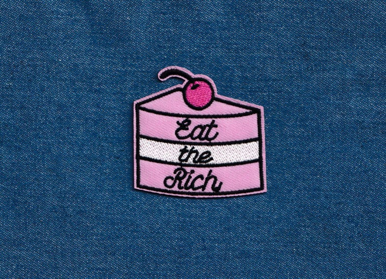 May include: Pink and white embroidered patch of a layered cake with a cherry on top. The text "Eat the Rich" is embroidered on the cake.